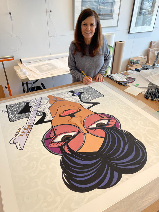 Pop artist Michelle Vella signing her portrait painting of The Artist formerly known as Prince with brown big eyes wearing big round sunglasses with a purple lens, black and purple hair, a black moustache, holding his guitar wearing a silver jacket with white and black writing of song titles and a white frilly shirt.