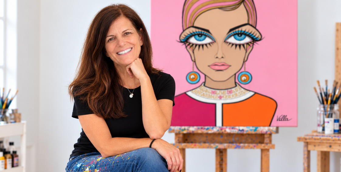 Artist Michelle Vella sits on a black rolling stool in a bright studio, smiling toward a large colorful portrait painting on an easel. The artwork shows a stylized woman with blue eyes, pink hair, and bright pink, orange, and white clothing.