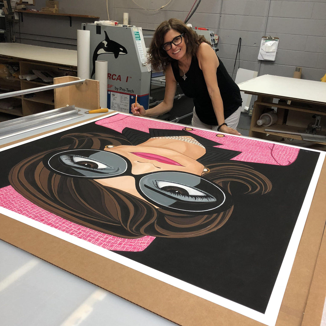 Artist Michelle Vella signing her Jackie Kennedy, Portrait painting and art print of Jackie Kennedy with brown big eyes wearing big round black sunglasses with hot pink lips, wearing a pearl necklace and earrings and a pink Chanel suit  and Chanel Pillbox hat on a black painted background